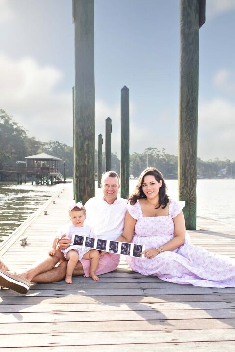 Family of three sitting on the dock at Church of the Cross in Bluffton, SC during pregnancy announcement session, holding ultrasound photos with the May River in the background