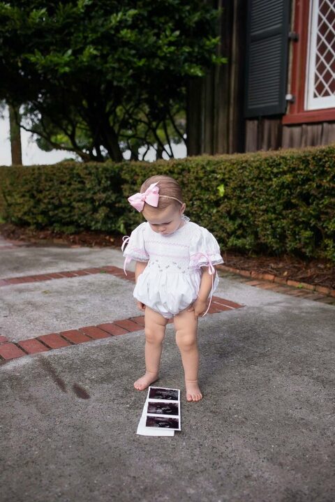 Little girl standing alone on walkway holding pregnancy ultrasound photos, wearing white outfit and bow during family announcement session in Bluffton, SC.