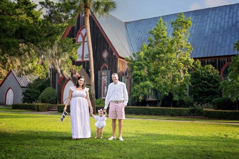 Family of three walking hand-in-hand on lawn in front of historic Church of the Cross in Bluffton, SC, mother holding ultrasound images announcing second pregnancy.