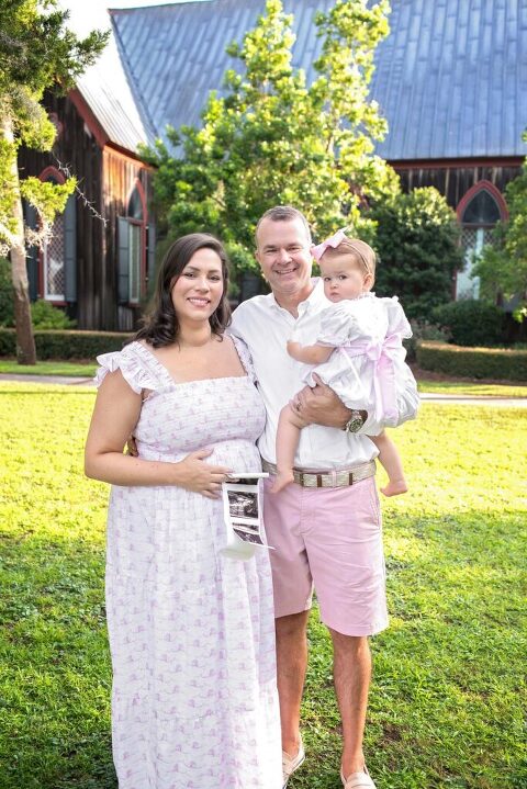 Expecting parents standing in front of Church of the Cross in Bluffton, SC holding toddler and ultrasound images for outdoor pregnancy announcement.