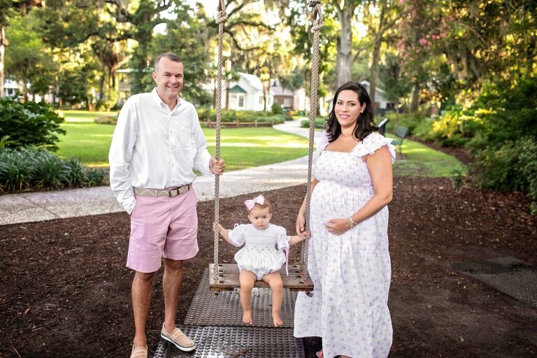 Parents sitting on brick steps smiling as toddler holds and examines ultrasound photos during outdoor pregnancy announcement session in Bluffton, SC.