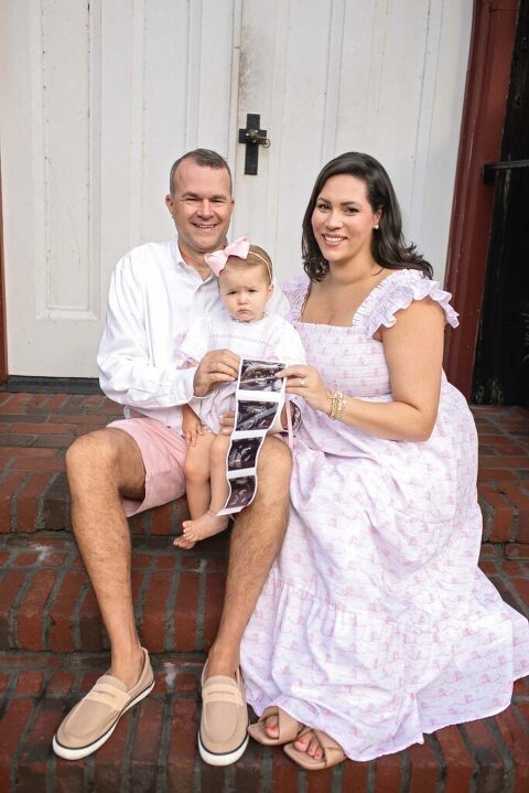 Parents sitting on brick steps holding toddler and ultrasound pictures during outdoor pregnancy announcement session at Church of the Cross in Bluffton, SC.