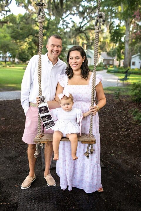 Family of three standing by a wooden swing under Spanish moss trees at Church of the Cross in Bluffton during pregnancy announcement session.