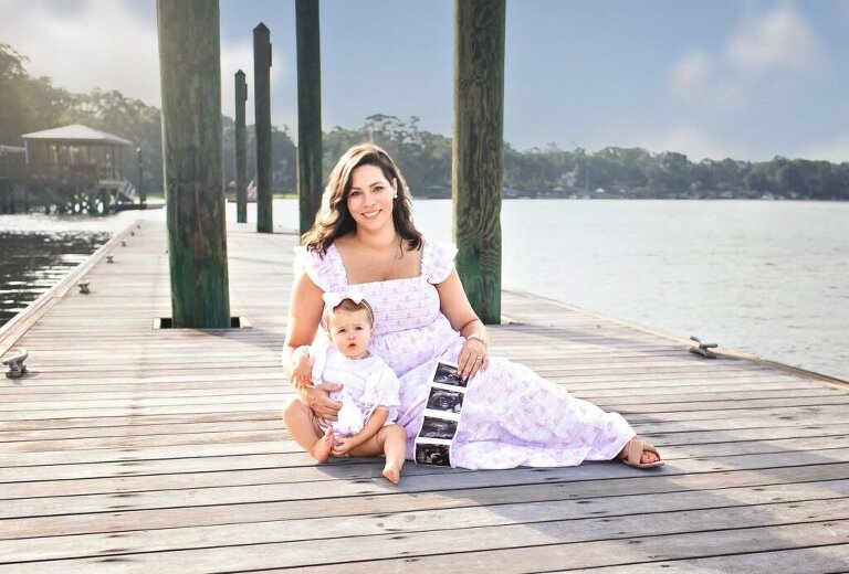 Mother and toddler sitting together on wooden dock in Bluffton, SC smiling and holding ultrasound photos to announce second pregnancy.