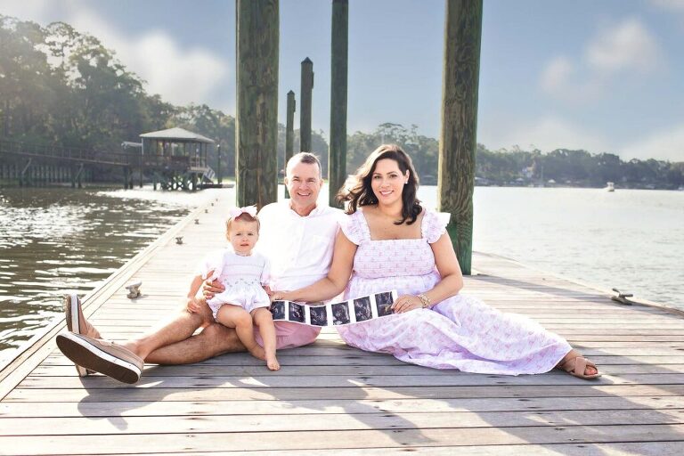 Family sitting on wooden dock in Bluffton, SC holding ultrasound strip to announce second pregnancy, with May River and dock pilings in the background.