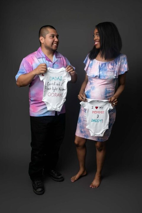 First-time parents standing together holding custom baby onesies and smiling during creative studio pregnancy announcement photoshoot in Beaufort, South Carolina.