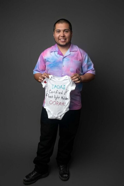 Expectant father smiling and holding custom baby onesie that reads “Dada’s Can’t Wait for His Little Oorah!” during studio pregnancy announcement session in Beaufort, SC.