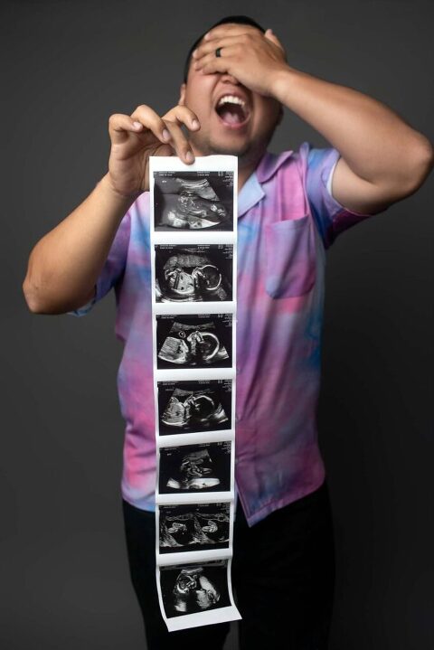 Expectant father laughing and covering his eyes while holding a long strip of ultrasound photos during a playful pregnancy announcement studio session in Beaufort, SC.