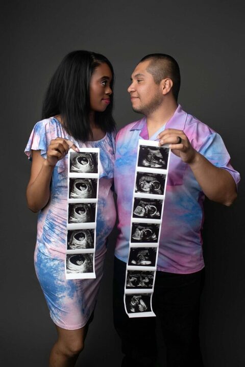 Couple smiling at each other and holding ultrasound strip during fun, modern pregnancy announcement photo session in photography studio in Beaufort, South Carolina.