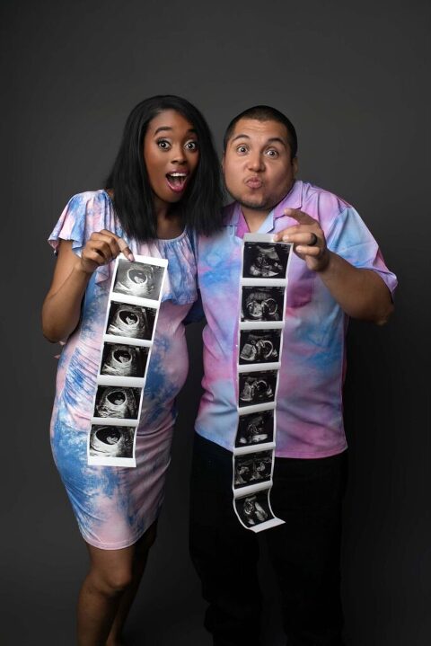 First-time parents in fun studio pregnancy announcement session holding a long strip of ultrasound images and reacting with surprised expressions, wearing matching pastel tie-dye outfits in Beaufort, SC.